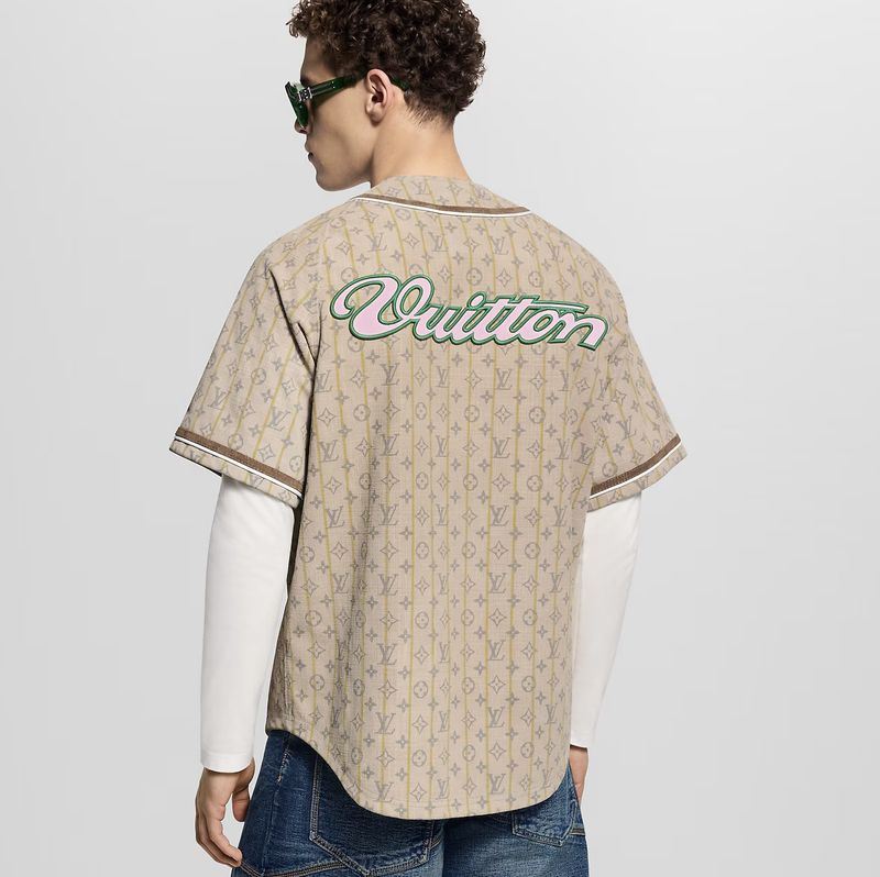 Man wearing beige Louis Vuitton logo baseball jersey with a long sleeve underneath and dark-wash jeans standing in a photo studio.