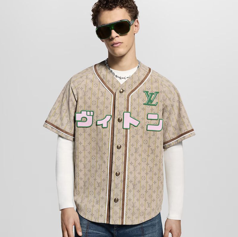 A stylish adult male poses indoors wearing a Louis Vuitton baseball jersey, jeans, and sunglasses, showcasing a modern fashion look.