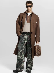 Model wearing dark sunglasses, brown coat, patterned trousers, and holding a checkered vanity case against a white background