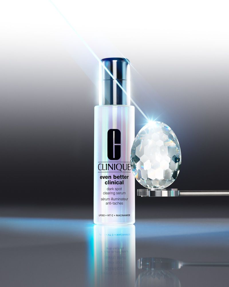 Studio shot of Clinique Even Better Clinical dark spot serum with a diamond-like glass orb, against a grey gradient background with blue light flares.
