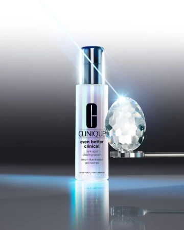 Studio shot of Clinique Even Better Clinical dark spot serum with a diamond-like glass orb, against a grey gradient background with blue light flares.