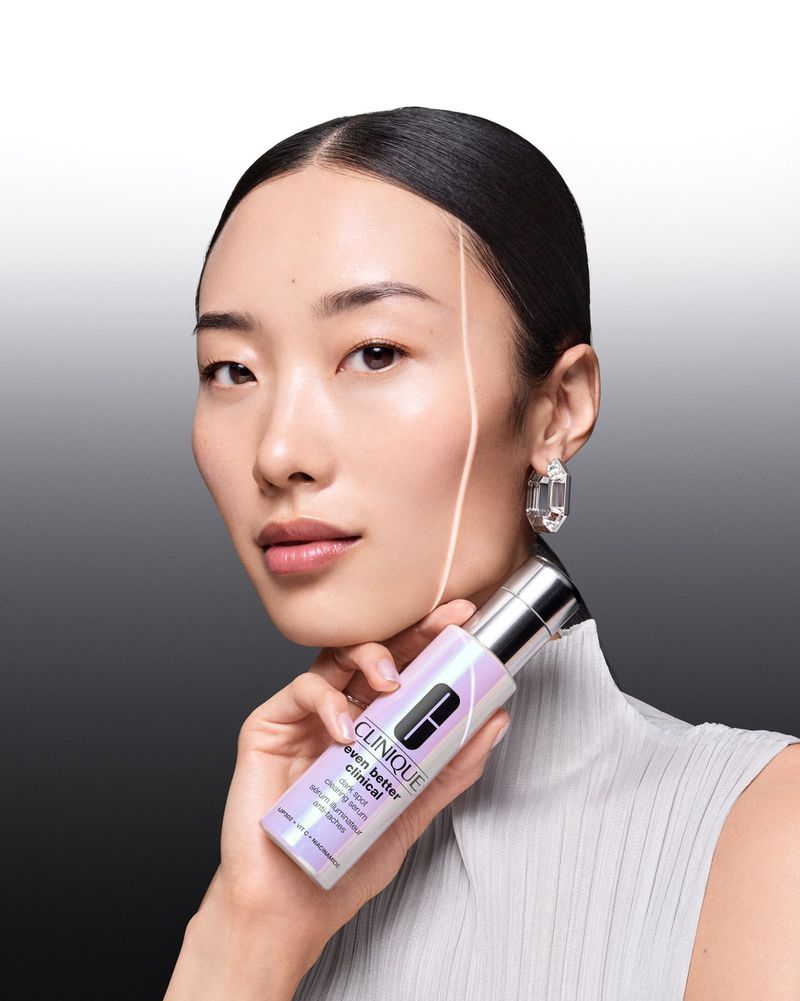 Portrait of an Asian woman holding a Clinique 'Even Better Clinical' serum bottle, wearing a light-colored textured top and large earrings.