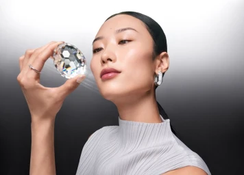 An East Asian woman holding a large faceted crystal reflecting light against a gradient grey studio backdrop.