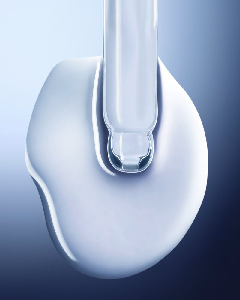 Close-up of a clear serum droplet with a glass dropper against a soft blue gradient background in a beauty or cosmetic product shot.