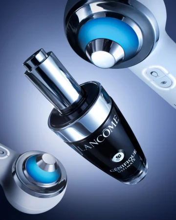 Close-up studio shot of Lancôme Génifique Ultimate serum bottle with chrome accents, next to two skin rejuvenation devices with blue lights.