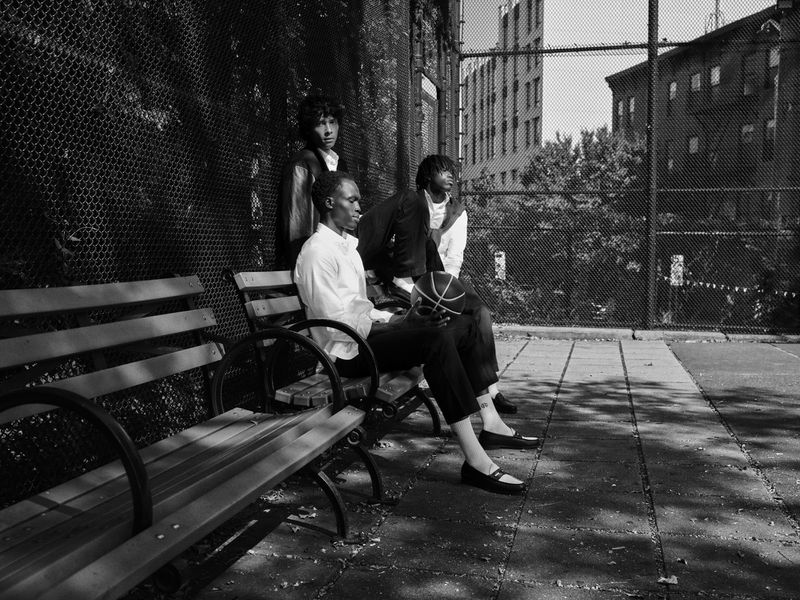 Three people, a basketball player among them, are sitting on park benches near a chain link fence with buildings in the background.