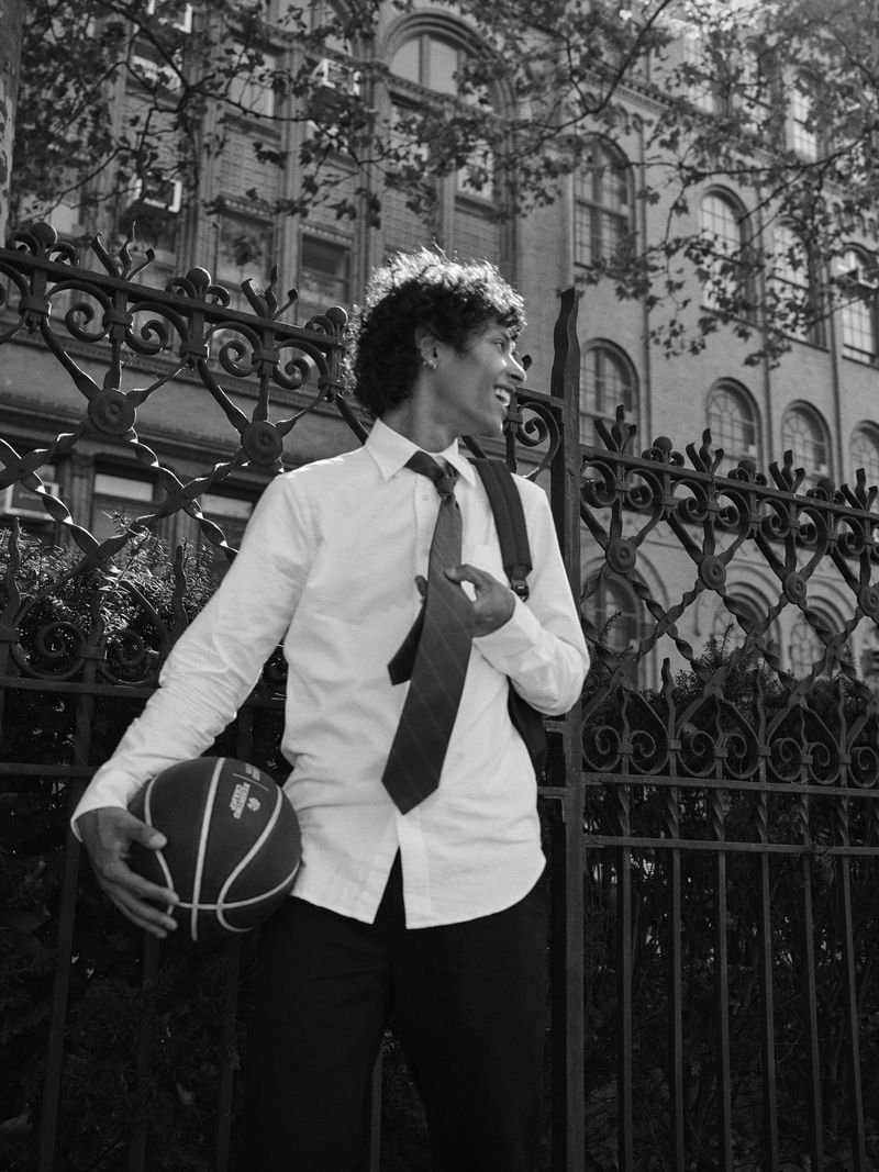 Black and white shot of an adult male in a dress shirt and tie holding a basketball in front of an ornate fence.