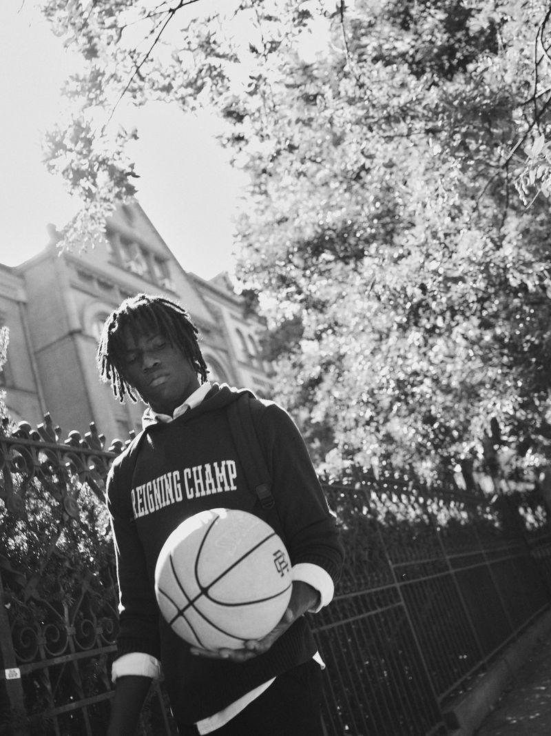 Monochromatic portrait of a young African American male holding a basketball in an urban setting with a building and leafy trees.