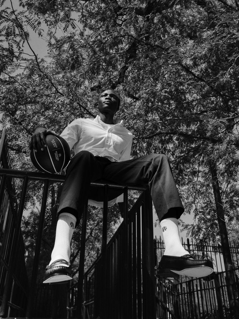 Black and white portrait of a young man outdoors sitting with a basketball against a tree backdrop in a low angle shot.