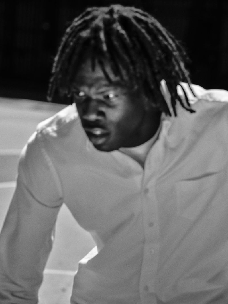 Black and white portrait of a Black adult male with dreadlocks wearing a white button-down shirt. He is in motion with a surprised expression.