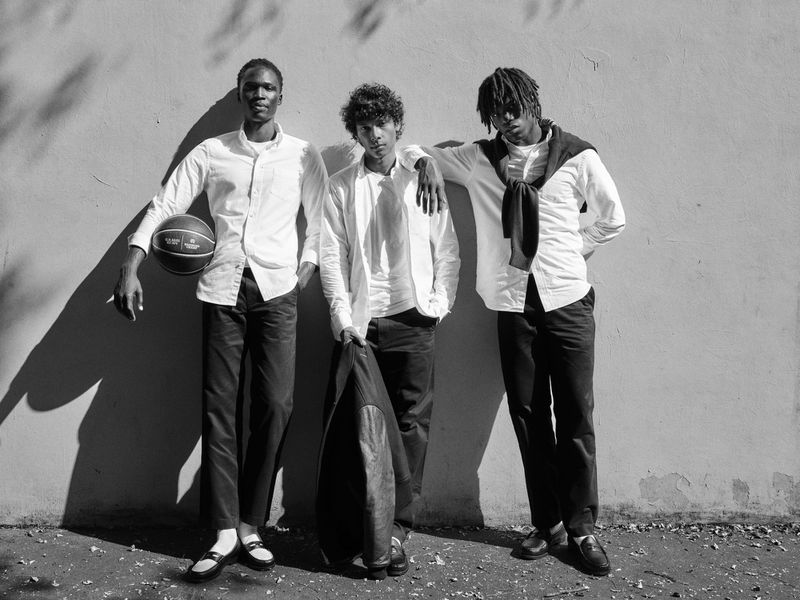 Black and white shot of three young men posing against a wall, one holding a basketball and another a jacket, in an outdoor setting.