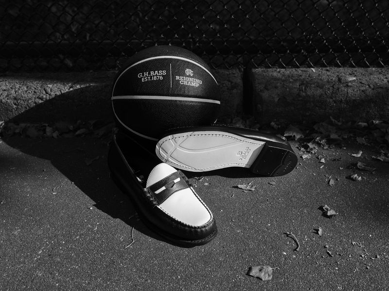Black and white photograph features G.H. Bass penny loafers with a basketball on an outdoor, concrete surface with a metal fence.
