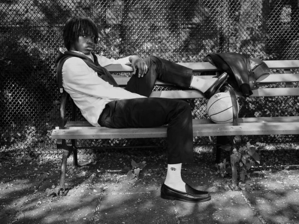Young man with dreadlocks seated on a park bench wearing loafers and a sweater draped over his shoulders
