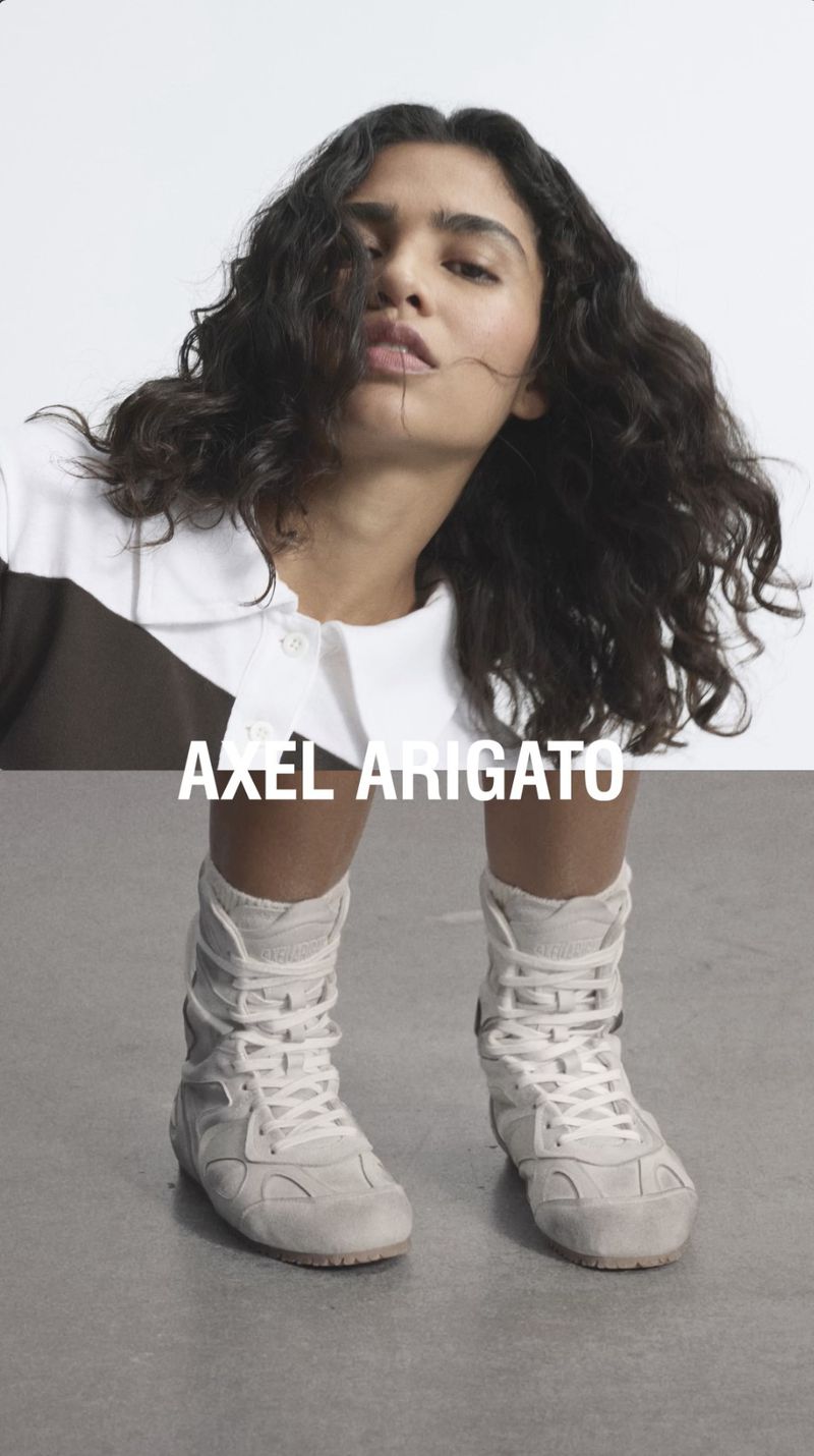 A young adult woman with curly dark hair poses wearing white tennis shoes and socks against a stark white studio backdrop.