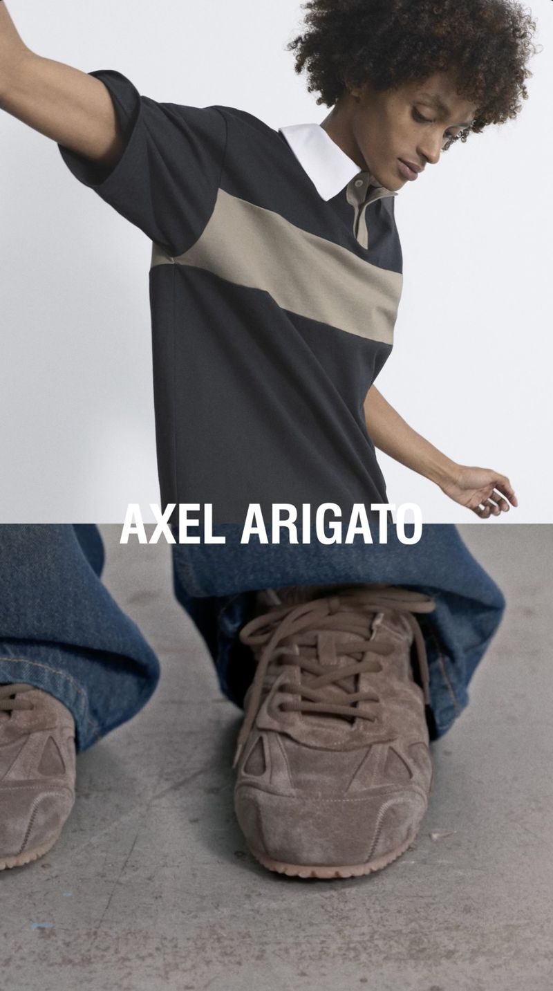A person wearing a black and beige striped polo shirt with blue jeans and brown sneakers poses indoors for a fashion shot.