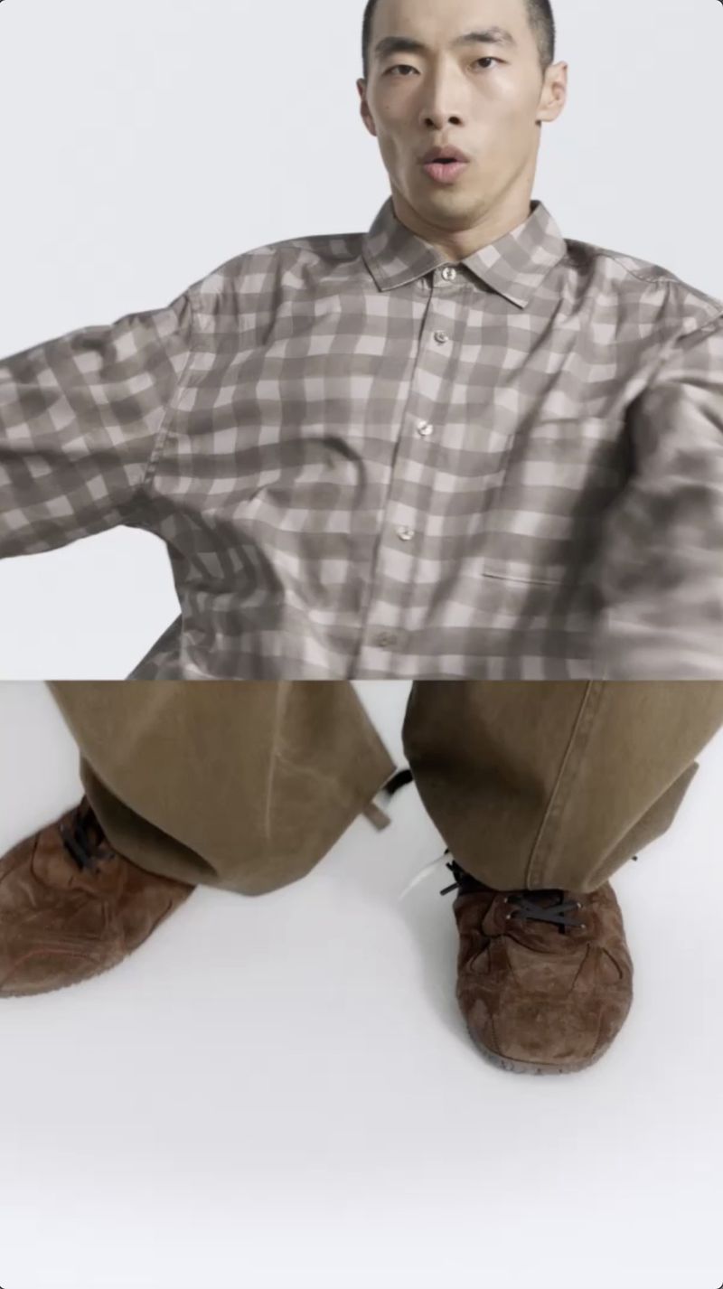 Asian man in a plaid shirt and brown trousers poses against a white backdrop, showcasing a casual and contemporary fashion look.
