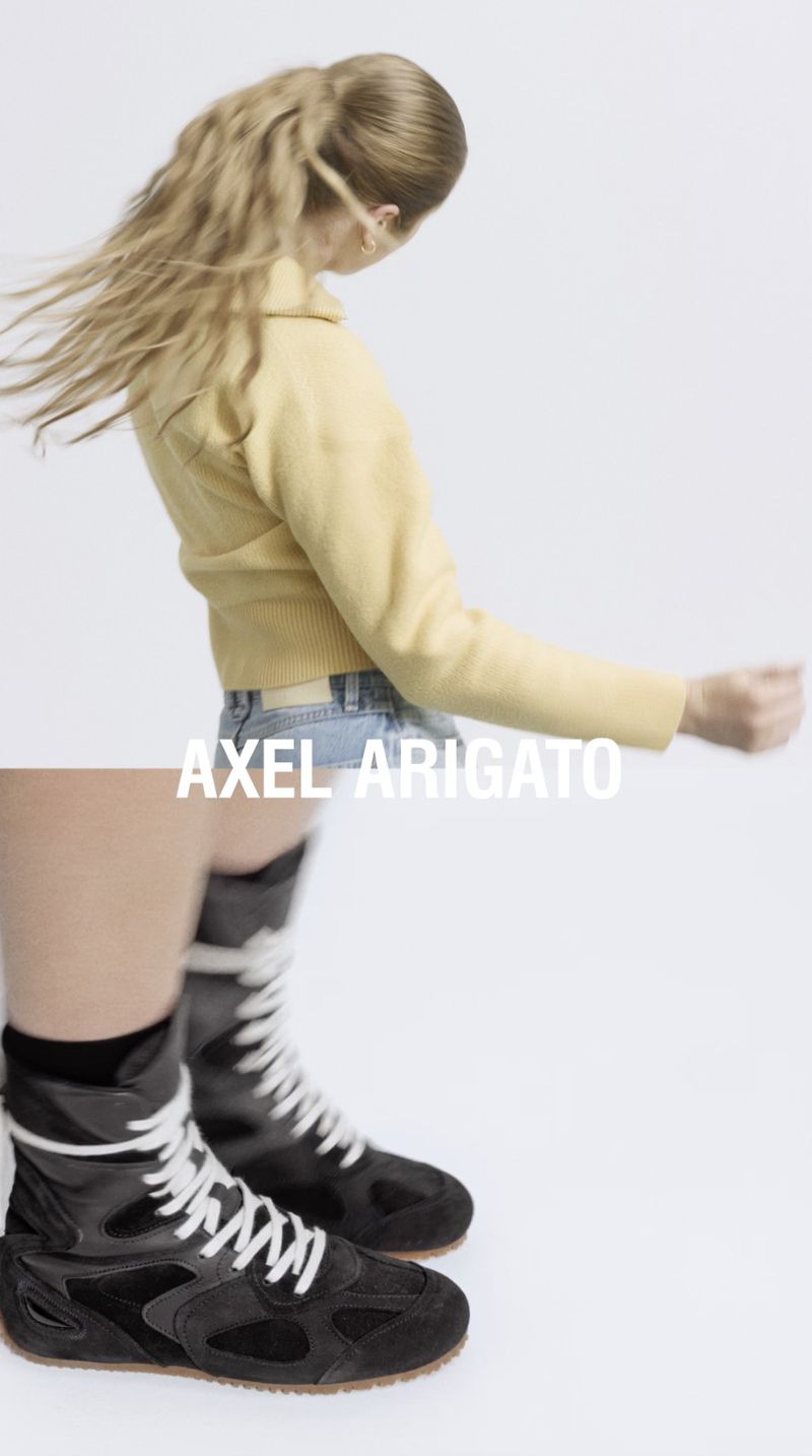 Fashion shot of a woman with blonde hair wearing a yellow jacket and jeans, paired with high-top black sneakers with white laces.