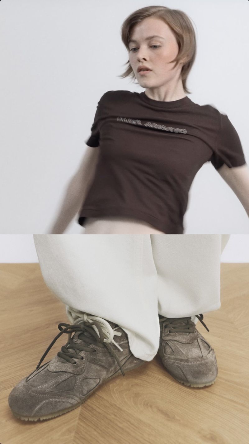 Fashion shot featuring a woman in a brown t-shirt and white pants with worn-out sneakers on a light wood surface and white background.