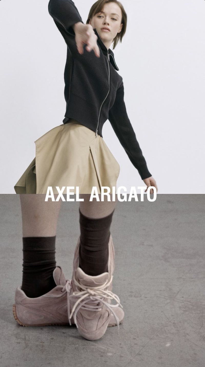 Fashion shoot features woman in black jacket and khaki skirt with Axel Arigato sneakers, striking a dynamic pose on a gray background.