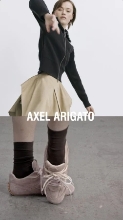 Fashion shoot features woman in black jacket and khaki skirt with Axel Arigato sneakers, striking a dynamic pose on a gray background.