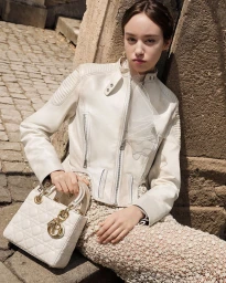 Model Camila Cainelli leaning against stonework wearing an off-white leather jacket and holding a white handbag for Dior