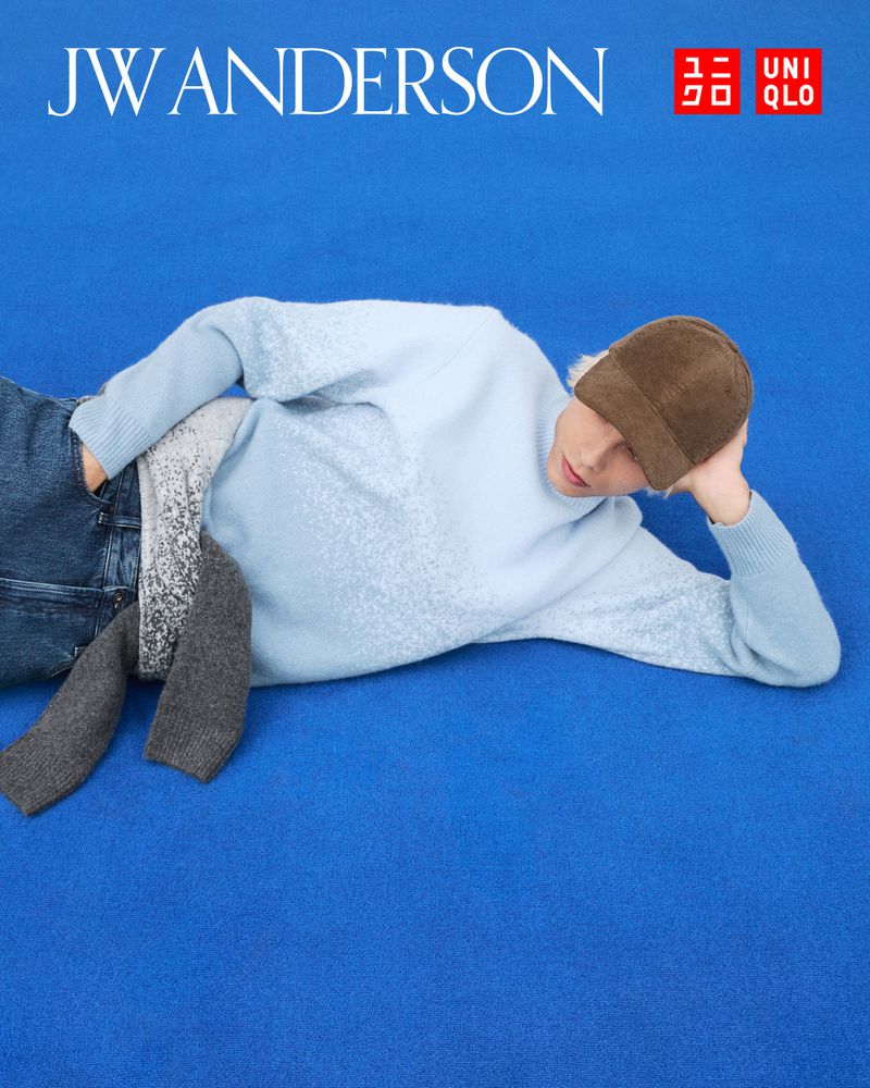 Fashion shot of person lying on a blue surface wearing jeans, light-blue sweater, and brown hat, advertising JW Anderson and Uniqlo.