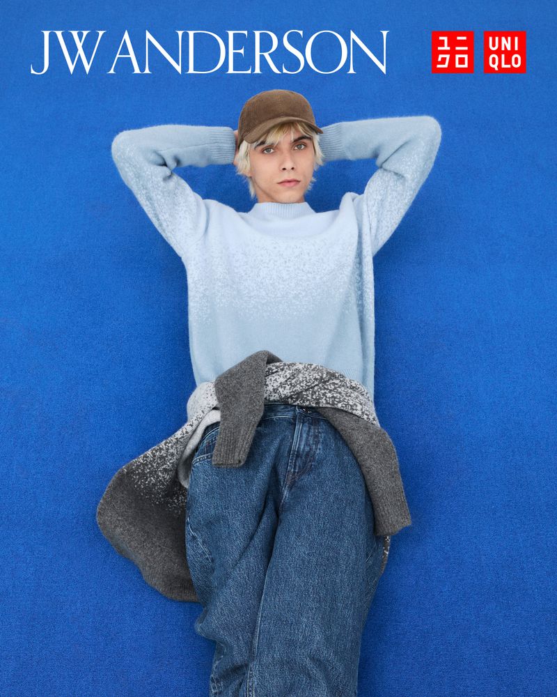 An androgynous model poses in a light blue sweater, jeans, and a brown cap against a blue backdrop, showcasing a modern, relaxed style.