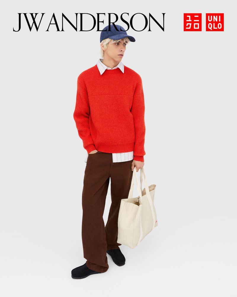 A blond male model wearing a red sweater, brown pants, black shoes and a navy hat poses with a tote bag in a fashion editorial shot.