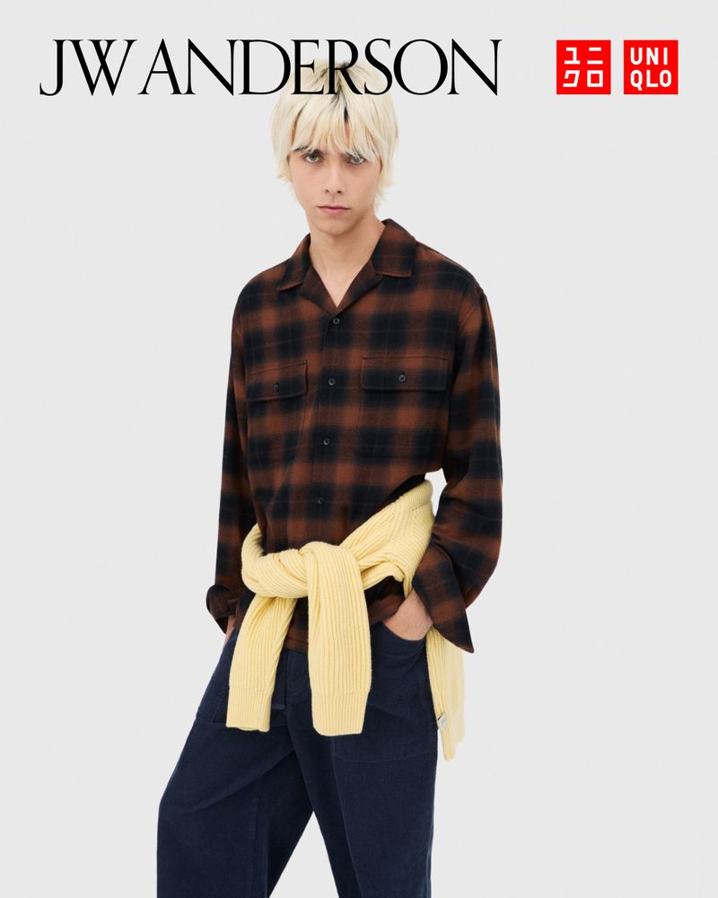 Man poses in a brown and black plaid shirt, navy pants, with a yellow sweater tied around his waist in a studio shot.