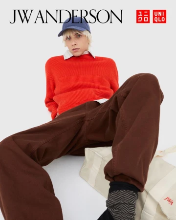Studio shot of a blond-haired adult wearing a blue baseball cap, orange sweater, and brown pants, sitting against a neutral backdrop.