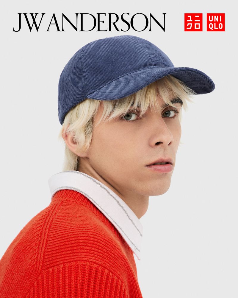 A young adult wears a corduroy baseball cap and an orange ribbed sweater with a white collared shirt against a light background.