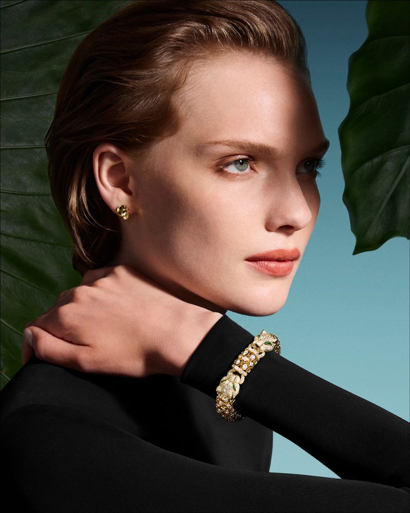 Close-up of a model wearing a bracelet and earrings. The jewelry is set against a background of tropical leaves and blue sky.
