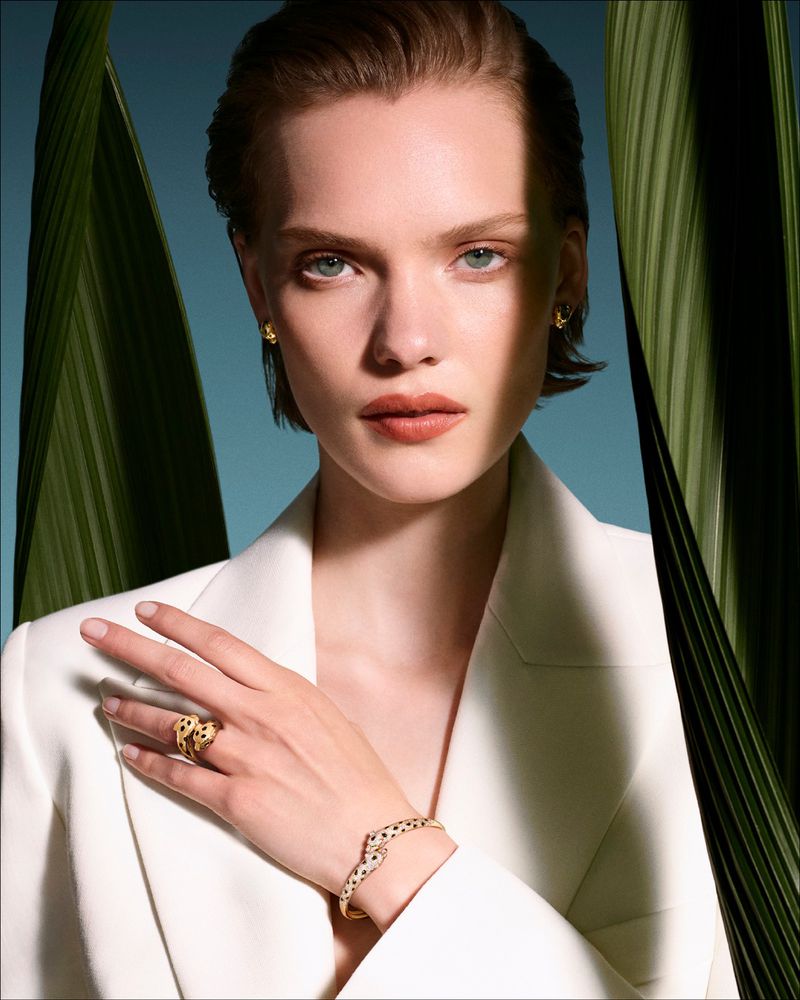 Model wearing a white blazer and gold panthère jewellery framed by large green palm leaves for Cartier