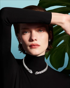 Model wearing a diamond panther necklace and black turtleneck against a light blue backdrop with tropical foliage.