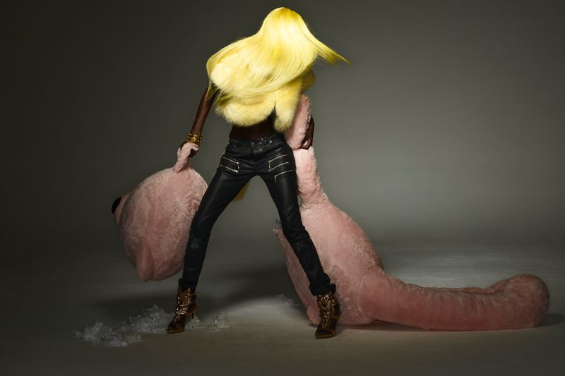 A black woman with blond hair and leather pants poses with a large pink teddy bear against a gray studio backdrop in a fashion shot.