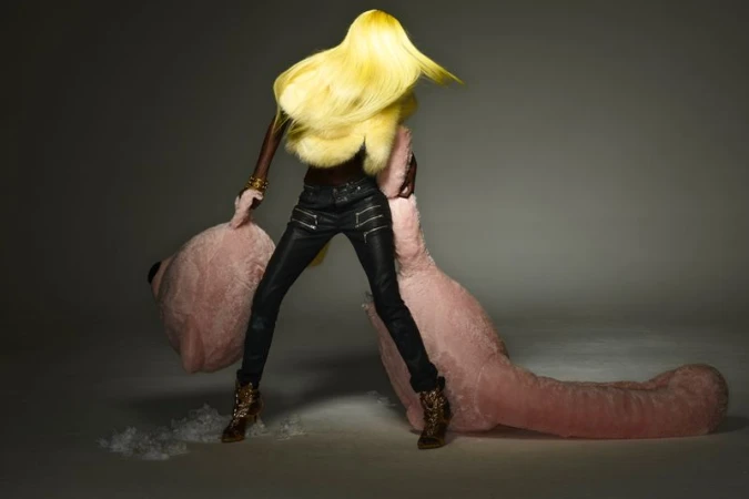 A black woman with blond hair and leather pants poses with a large pink teddy bear against a gray studio backdrop in a fashion shot.