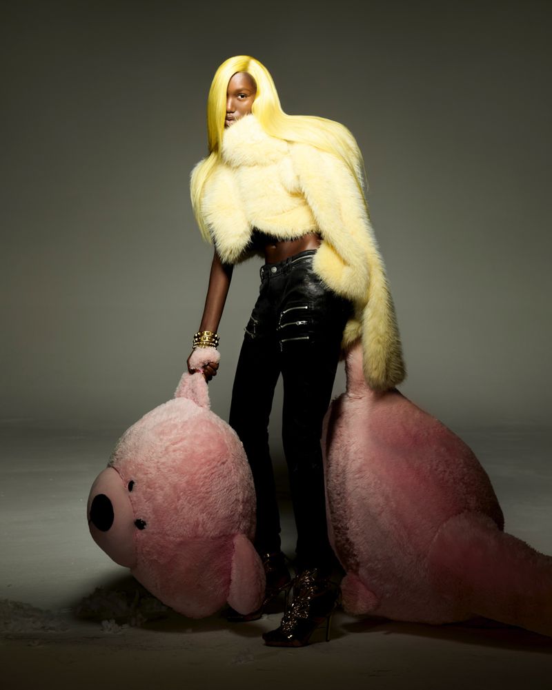 A woman with yellow hair wearing a light yellow fur jacket, black pants, and black wedge shoes poses with two pink teddy bears.