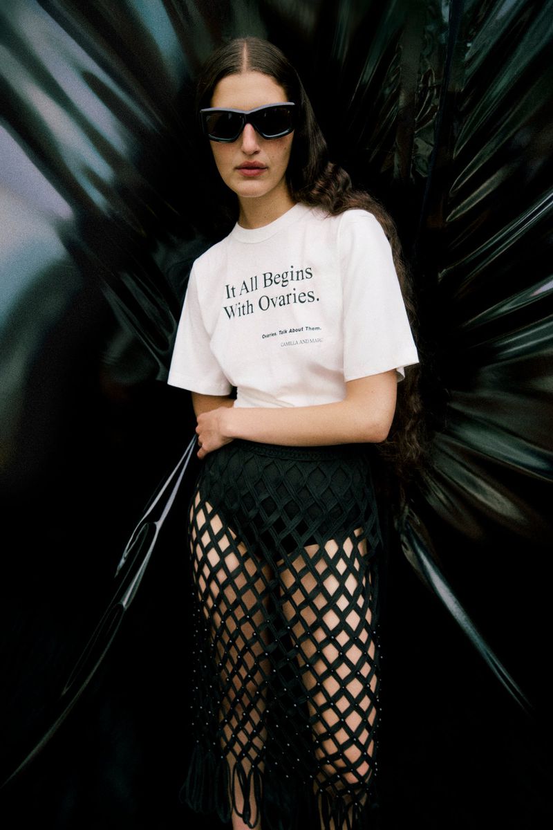 A young woman poses in a white t-shirt, net skirt, and sunglasses, standing in front of a black backdrop, fashion portrait.