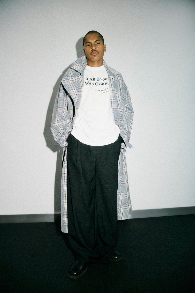 Fashionable adult man in a plaid coat and black pants poses in front of a neutral background in a studio, looking at the camera.