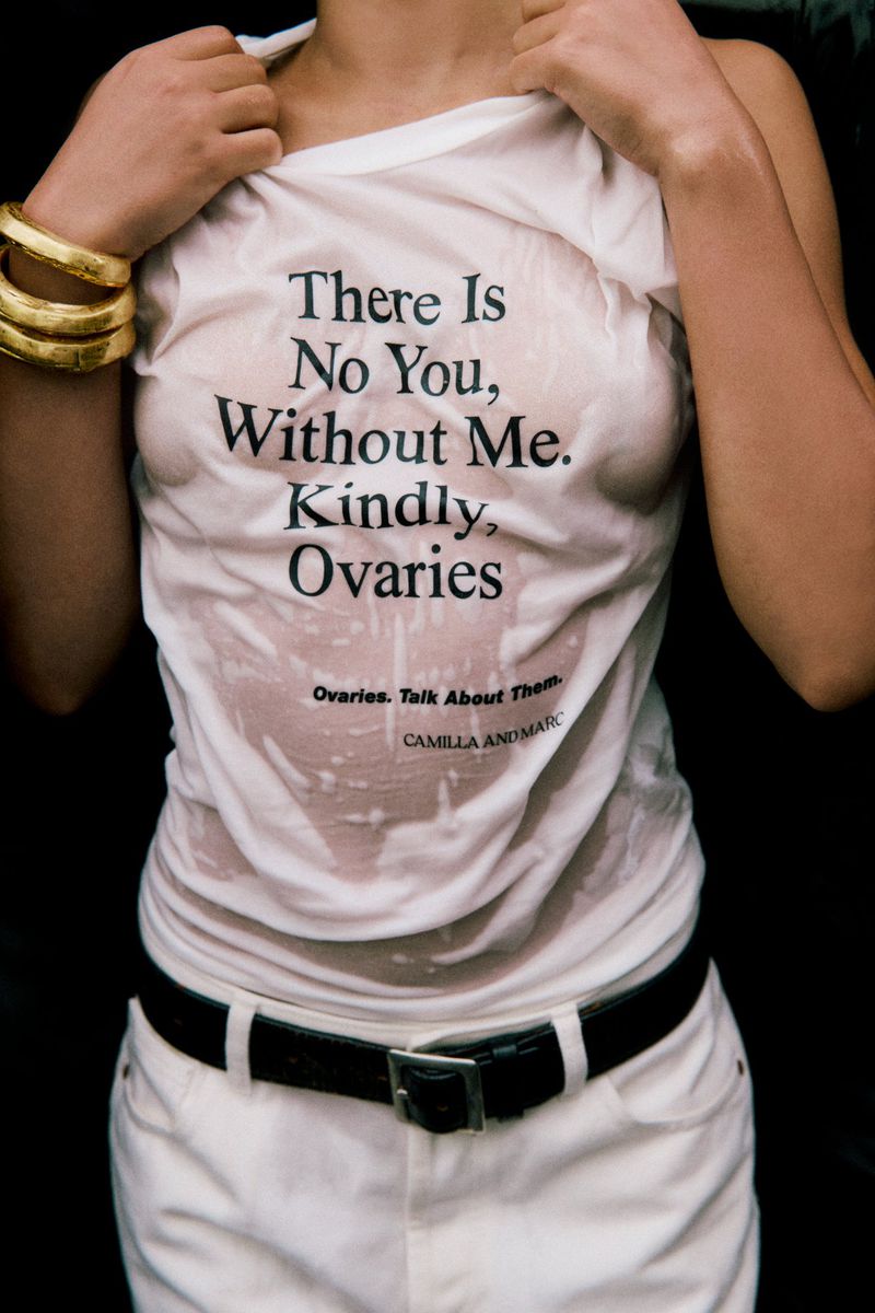 Close-up of a woman wearing a wet t-shirt with text, white jeans, and gold bangles. The black text on the shirt says, 'There Is No You, Without Me.'