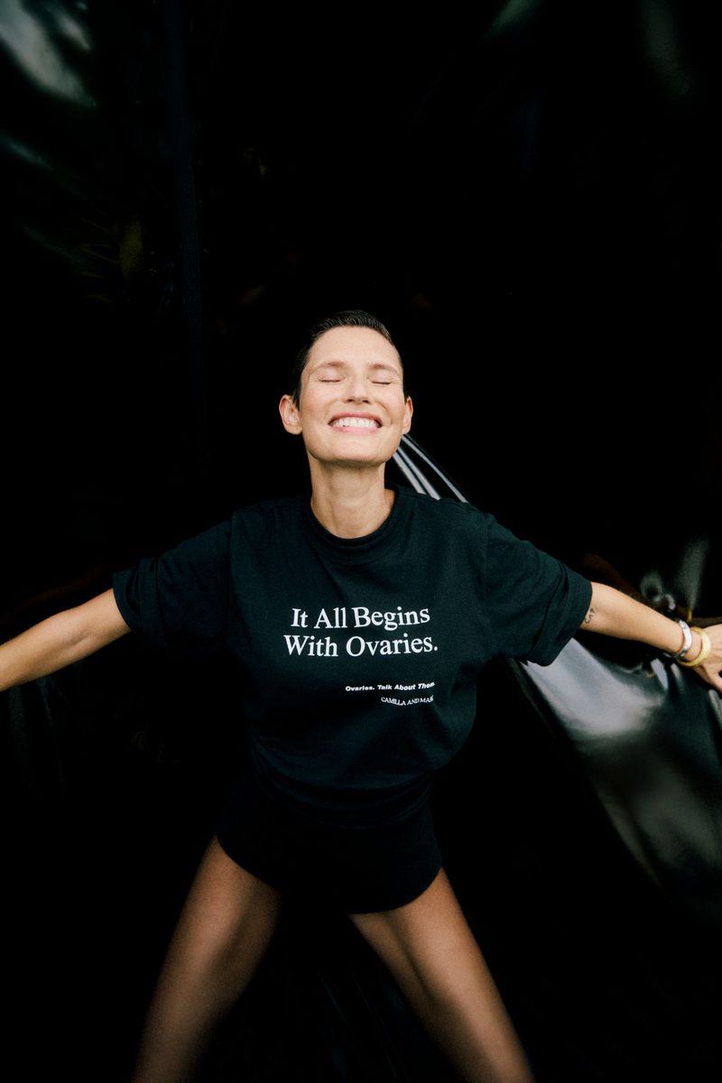 Woman in black t-shirt with empowering slogan smiles with eyes closed against a dark backdrop, arms slightly outstretched, indoor setting.