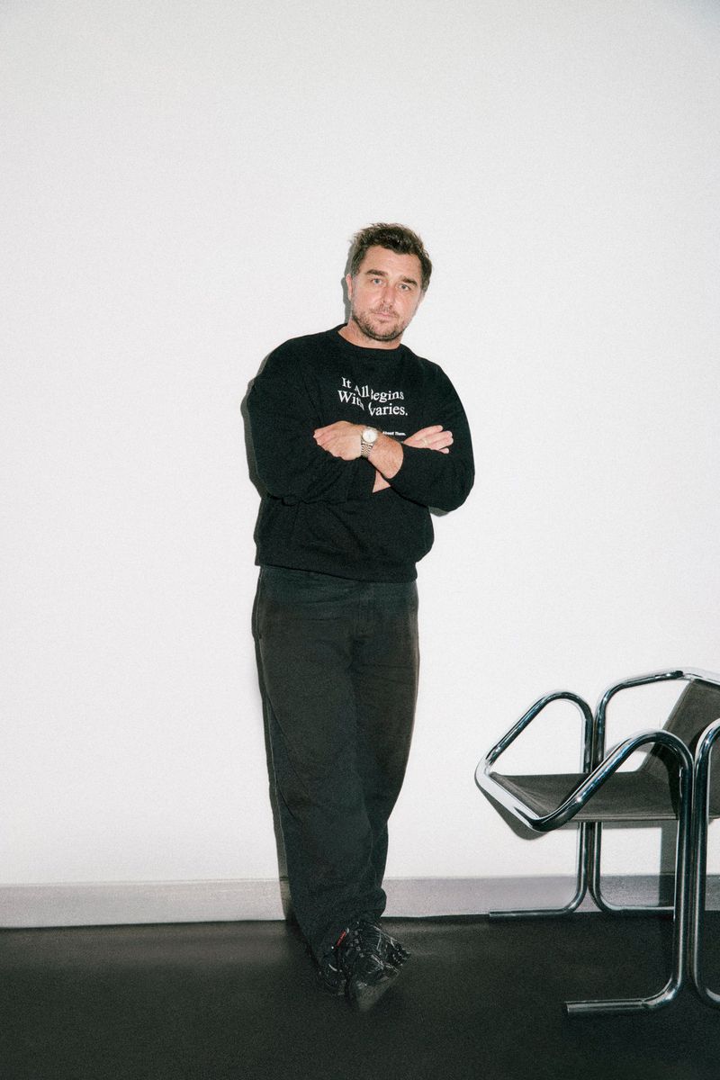 Man in a black sweatshirt standing in front of a white wall with his arms crossed, wearing dark pants and shoes, indoors.