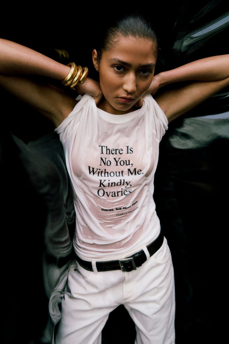 Woman in wet t-shirt with text, white pants, and gold bracelets poses against a dark background, high-angle view, beauty advertisement.