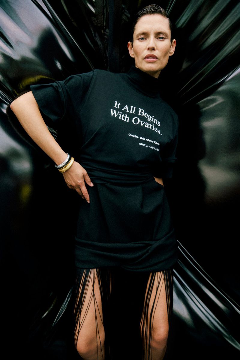 Woman in black dress with text poses against dark background. The dress has fringe detail. The woman has bracelets on her wrist.