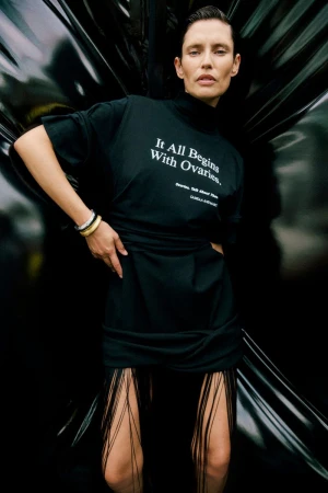 Woman in black dress with text poses against dark background. The dress has fringe detail. The woman has bracelets on her wrist.