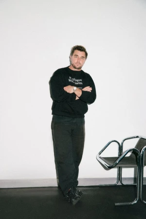Man standing with arms crossed wearing a black sweatshirt and dark trousers against a white wall next to a modern chair