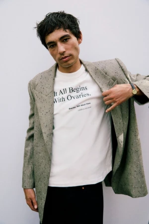 Man wearing a printed white t-shirt and speckled grey blazer against a white wall for Camilla and Marc.