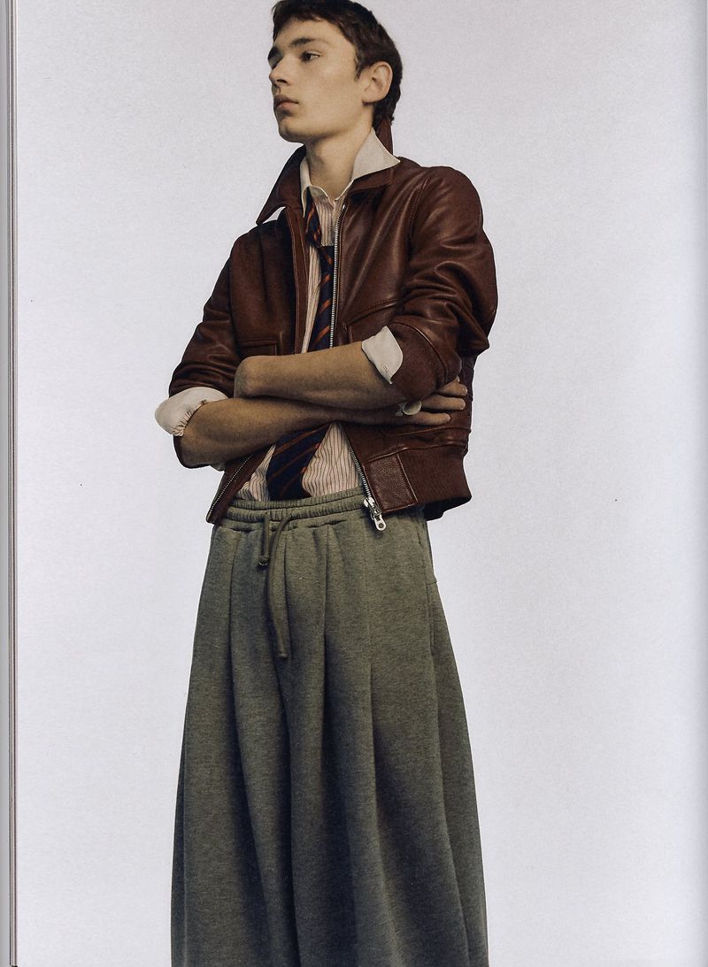 Male fashion model poses indoors wearing a leather jacket, collared shirt, tie, and oversized sweatpants against a white background.
