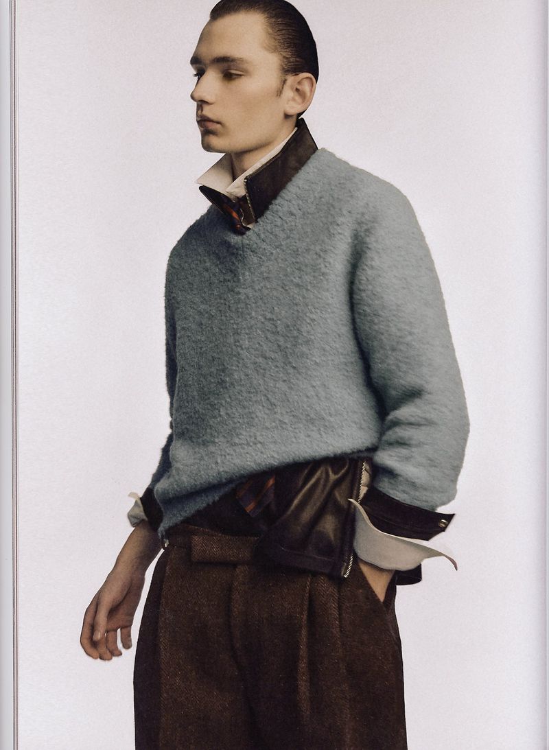 Man in light blue sweater, brown trousers, and white collared shirt. Standing indoors with a neutral backdrop, looking off to the side.