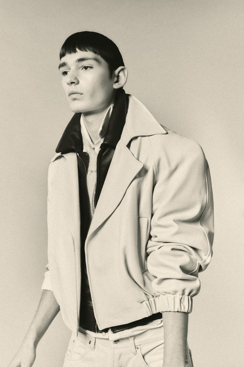 Portrait of a young adult with a bowl cut, wearing a light-colored jacket, collared shirt, and jeans against a neutral background.